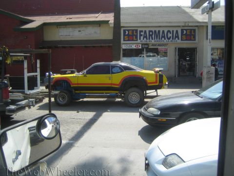 1990 Ford Bronco - yellow and red lifted custom coupe on big tires sits at a street curb in front of a Farmacia Del Sol pharmacy, seen past a side mirror.