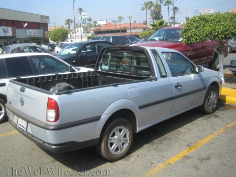 1990 Ford Bronco - Rear-quarter view of a silver Volkswagen Pointer single-cab pickup parked in a busy lot with shops and palm trees behind.