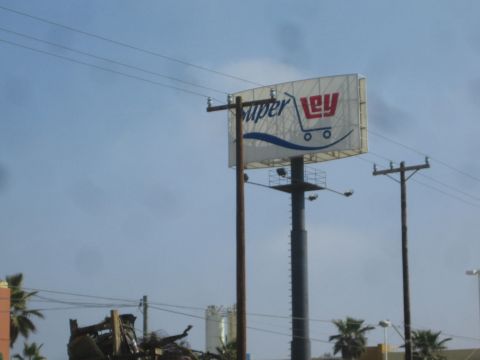 1990 Ford Bronco - tall Super Ley supermarket billboard with shopping-cart logo stands on poles beside power lines and palm trees under a hazy sky.