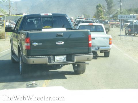 1990 Ford Bronco - dark green crew-cab pickup and a silver truck sit in slow traffic on a dusty rural highway near a Cruce Escolar school-crossing sign.