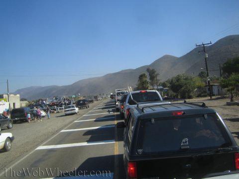 1990 Ford Bronco - long line of pickups and SUVs is parked along a two-lane rural road at the base of barren brown mountains under a clear sky.