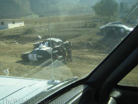 1990 Ford Bronco - white-and-blue police car number 08154 and a wrecked off-road vehicle sit in a dusty dirt lot, viewed through a vehicle windshield.