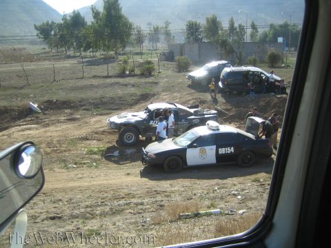 1990 Ford Bronco - black-and-white police car number 08154 parks beside an off-road race truck and people in a dusty field, seen through a car window.