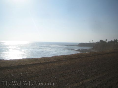 1990 Ford Bronco - Bright sun glints off the ocean along a rocky, gravelly coastline with low headlands in the distance under a clear blue sky.