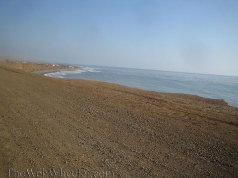 1990 Ford Bronco - wide gravel-covered coastal bluff slopes toward the ocean, with distant buildings and surf along a hazy shoreline.