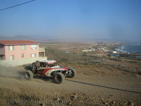 1990 Ford Bronco - red and white off-road race buggy speeds along a dusty dirt road on a bluff overlooking a coastal town and the ocean.