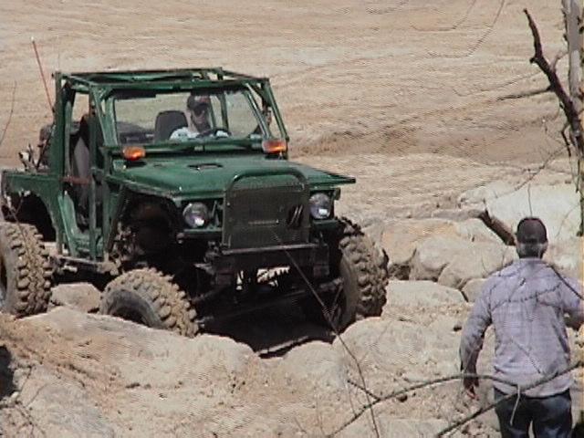 green off-road vehicle with large tires navigating rocky terrain, person watching