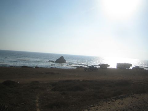 1990 Ford Bronco - RVs, trailers and vehicles are gathered on a sandy beachfront beside rocky outcrops as the sun glares brightly over the ocean.