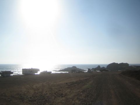 1990 Ford Bronco - wide dirt road leads toward RVs and vehicles parked along a rocky beachfront, with bright sun reflecting off the ocean.
