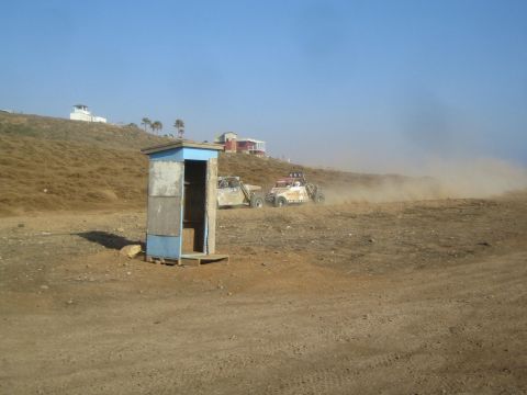1990 Ford Bronco - Two off-road race buggies kick up dust on a barren dirt course beside a small blue roadside outhouse, with houses on the hill behind.