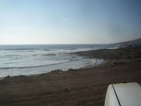 1990 Ford Bronco - Waves roll onto a rugged coastal beach with arid hills in the distance, viewed past the edge of a white vehicle hood.