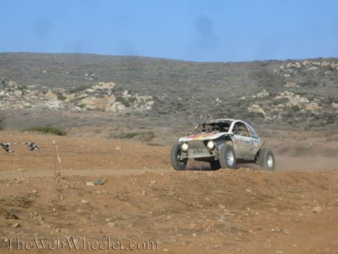 1990 Ford Bronco - white single-seat off-road race buggy kicks up dust as it races over desert dirt below scrub-covered hills.
