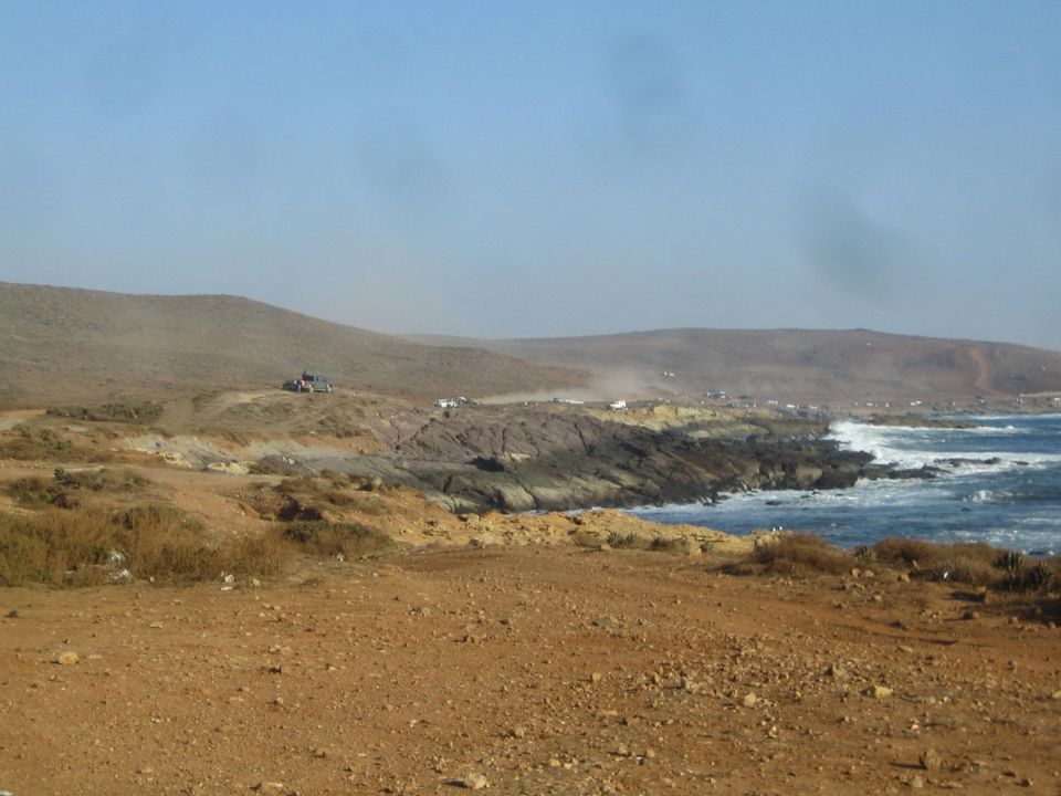 Arid coastal landscape with rocky shoreline, dust, and distant vehicles