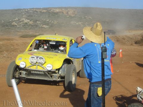 1990 Ford Bronco - man in a straw hat and blue hoodie holds a phone toward a yellow number 500 off-road race buggy at a dusty desert checkpoint.