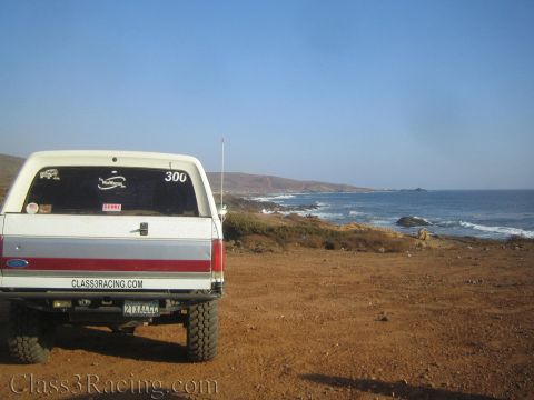 1990 Ford Bronco - tailgate of a white lifted pickup with race decals faces a rugged coastal bay under a hazy sky.