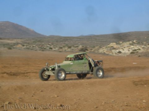 1990 Ford Bronco - green tube-frame off-road buggy speeds across desert dirt, trailing dust below distant arid hills.