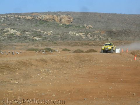 1990 Ford Bronco - yellow off-road race truck travels a distant dirt course marked by cones and buried tires below scrub-covered hills.