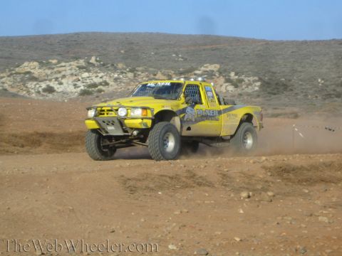 1990 Ford Bronco - yellow off-road race truck with light bars charges over a dirt rise, throwing up dust against arid hills.