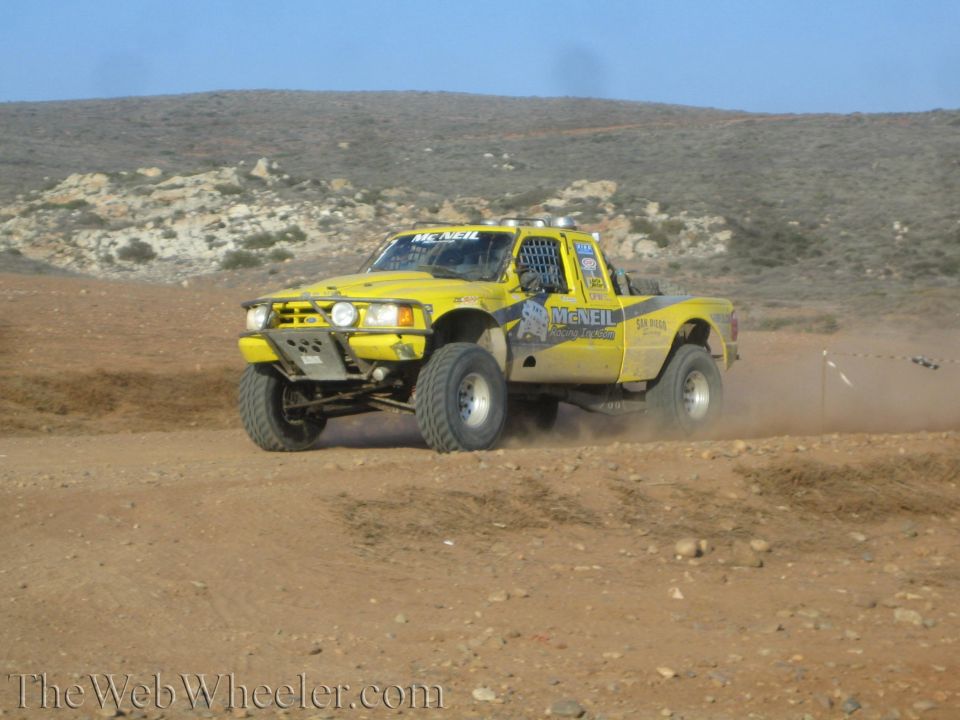 Yellow off-road race truck launching over a dirt rise, kicking up dust