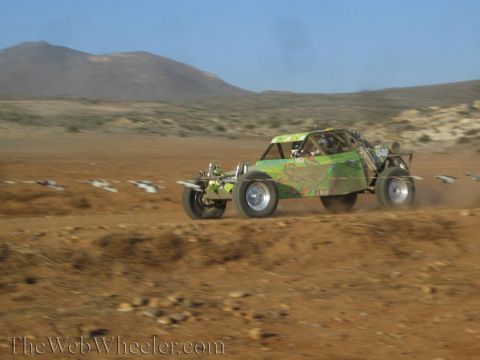 1990 Ford Bronco - green camo-painted tube-frame off-road buggy races across desert dirt, motion-blurred and trailing dust.