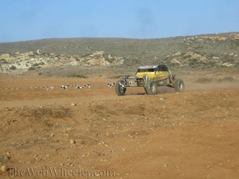 1990 Ford Bronco - yellow off-road buggy speeds along a desert dirt track edged with buried tires below scrub-covered hills.