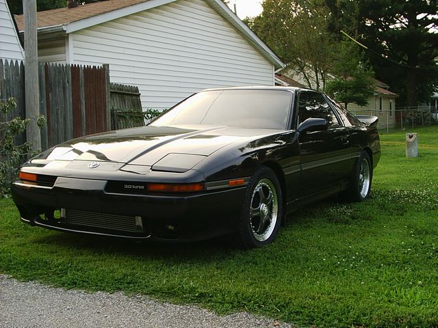 Black coupe with pop-up headlights and '3.0 TURBO' badge, parked on grass