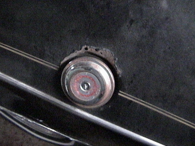 Close-up of a circular car emblem with red and silver accents, mounted on a dark surface with some rust around the edges