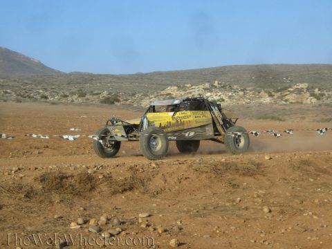 1990 Ford Bronco - yellow tube-frame off-road race buggy speeds across desert dirt past course markers, trailing a plume of dust.