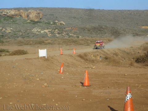 1990 Ford Bronco - distant off-road vehicle kicks up dust on a winding desert course lined with orange cones and a white sign board.