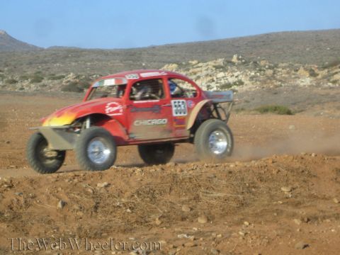 1990 Ford Bronco - red Beetle-based Baja Bug with flame graphics races across desert dirt, kicking up dust below arid hills.