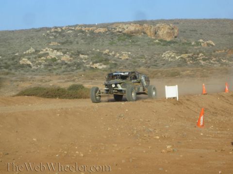 1990 Ford Bronco - off-road race buggy crests a dirt berm on a desert course marked with orange cones and a white sign board.