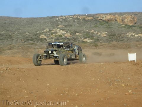 1990 Ford Bronco - dark-colored tube-frame off-road buggy races across desert dirt below a rocky scrub hillside, trailing dust.