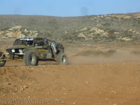 1990 Ford Bronco - Off-road race vehicles, including a number 133, churn through dust atop a desert dirt berm below scrub-covered hills.
