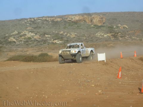 1990 Ford Bronco - white off-road race truck with light bars travels a coned desert dirt course, trailing dust below a rocky hillside.