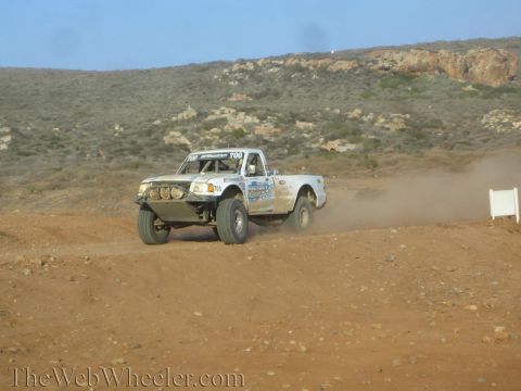 1990 Ford Bronco - white number 700 off-road race truck with light bars speeds across desert dirt, kicking up dust below arid hills.