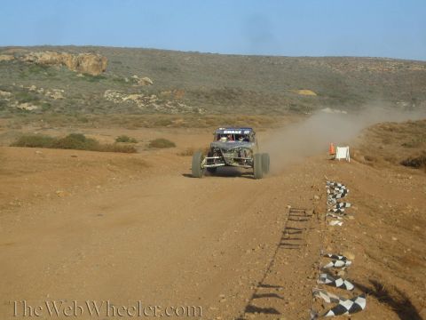 1990 Ford Bronco - off-road race buggy speeds away down a desert dirt track lined with checkered-flag markers, trailing dust.