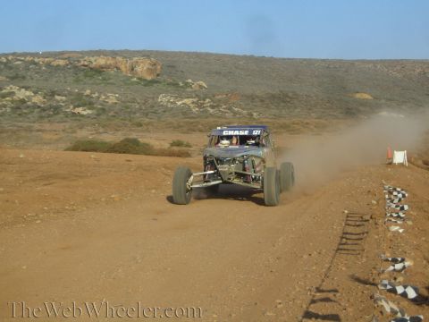 1990 Ford Bronco - blue and dark off-road buggy marked Chase 121 races up a desert dirt track lined with checkered-flag markers.