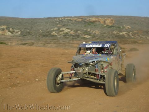 1990 Ford Bronco - Chase 121 tube-frame off-road buggy charges toward the camera across desert dirt with coilover shocks visible.