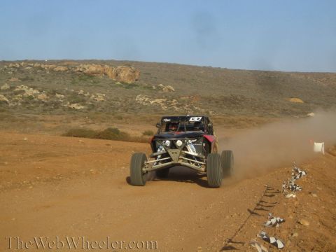 1990 Ford Bronco - off-road race buggy speeds away up a desert dirt track lined with checkered markers, trailing dust below arid hills.