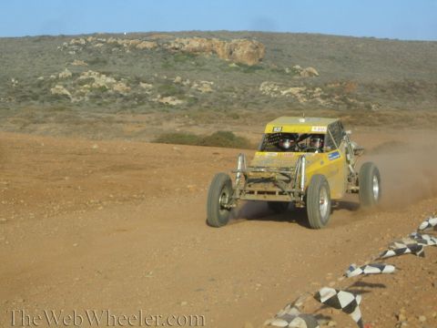 1990 Ford Bronco - yellow tube-frame off-road buggy races along a desert dirt track lined with checkered-flag markers below scrub hills.