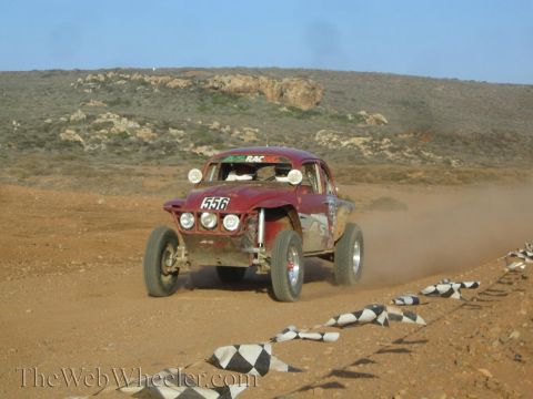 1990 Ford Bronco - weathered dark-red Beetle-based Baja Bug numbered 556 races along a desert track lined with checkered markers.
