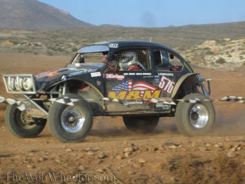 1990 Ford Bronco - black Beetle-based Baja Bug numbered 576 with M&M graphics races across desert dirt, motion-blurred and trailing dust.