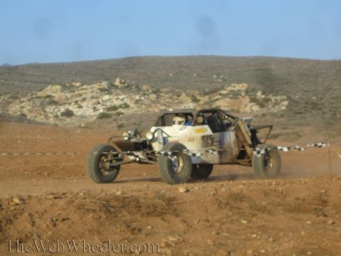 1990 Ford Bronco - white tube-frame off-road race buggy speeds across a dusty desert course past a checkered course marker, with brown hills behind.
