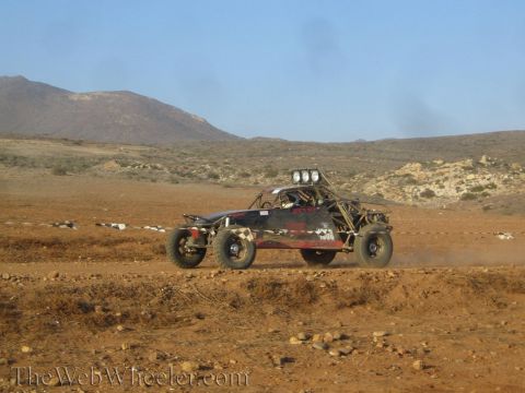 1990 Ford Bronco - dark tube-frame off-road buggy with a roof-mounted light bar races across reddish desert terrain trailing dust.