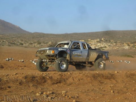 1990 Ford Bronco - gray off-road race truck with BFGoodrich and Thing Motorsports decals catches air over rocky desert ground.