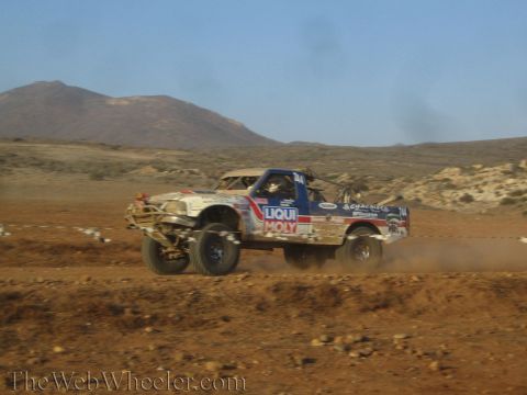 1990 Ford Bronco - white and blue off-road race truck branded Liqui Moly powers through a dirt berm kicking up dust in the desert.