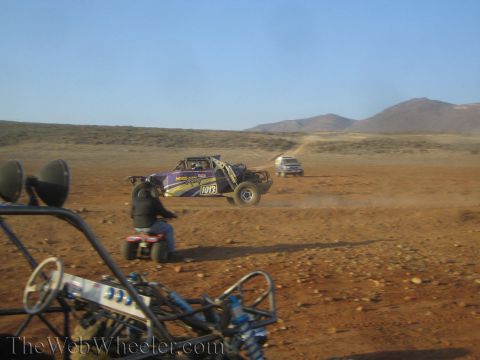 1990 Ford Bronco - purple off-road race buggy numbered 1015 jumps a desert rise while a spectator on a quad and a parked buggy watch in the foreground.