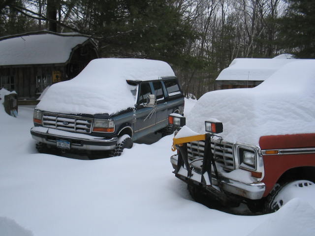 Dark blue SUV with snow plow attachment in front, parked next to a red vehicle covered in snow
