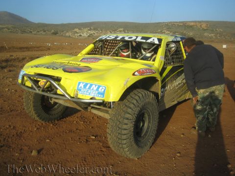 1990 Ford Bronco - yellow off-road race truck lettered Pistola sits at a desert staging area with King shocks decals as a crew member stands beside it.