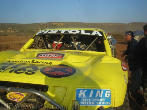 1990 Ford Bronco - Close rear view of a yellow off-road race truck lettered Pistola with King Off-Road Racing Shocks and American Racing decals, crew nearby.